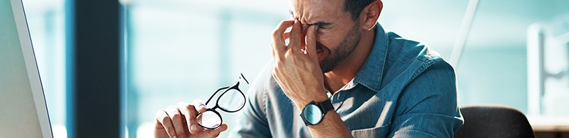 Mann sitzt vor einem Computerbildschirm und reibt sich die Augen, in der anderen Hand hält er eine Brille