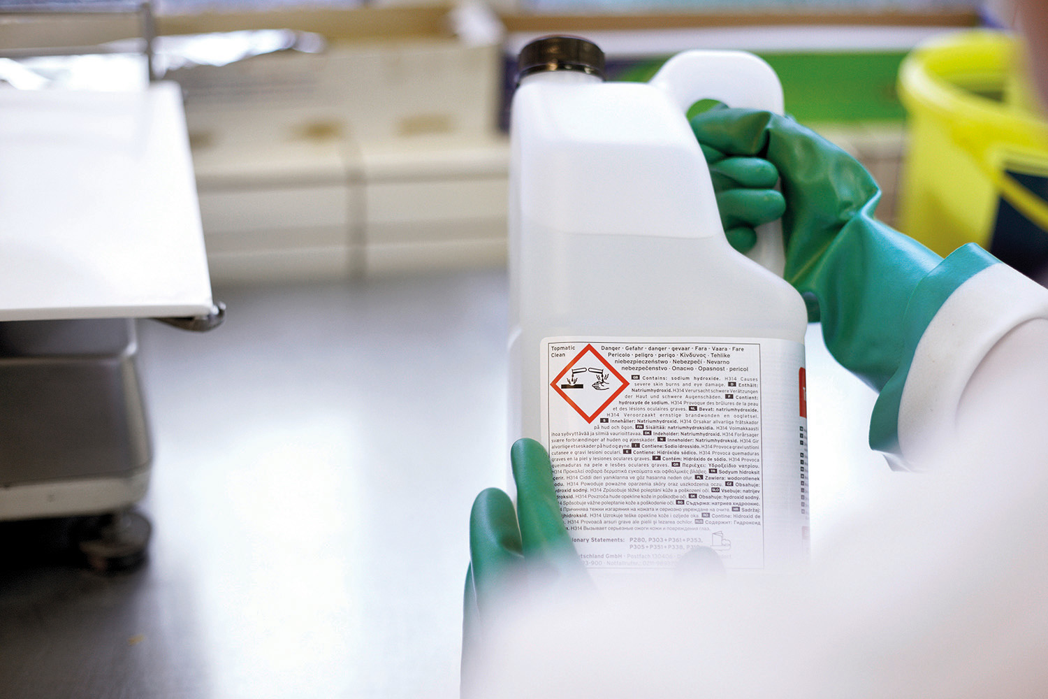 Person mit grünen Schutzhandschuhen hält in einem Labor eine weiße Chemikalienflasche mit Gefahrenpiktogramm (ätzend) auf dem Etikett.